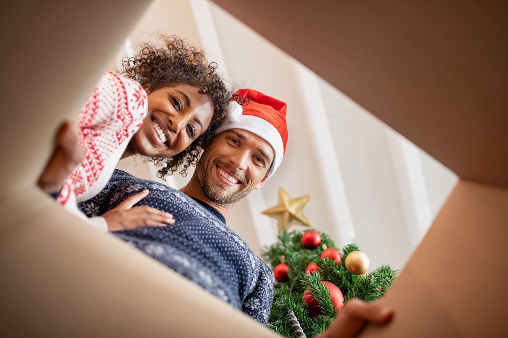 Heureux couple multiethnique embrassant tout en regardant à l'intérieur d'une boîte-cadeau. Jeune homme et belle femme africaine déballage cadeau de Noël avec arbre de Noël. Souriant jeune homme portant chapeau de père Noël avec femme regardant la caméra lors de l'ouverture présente.