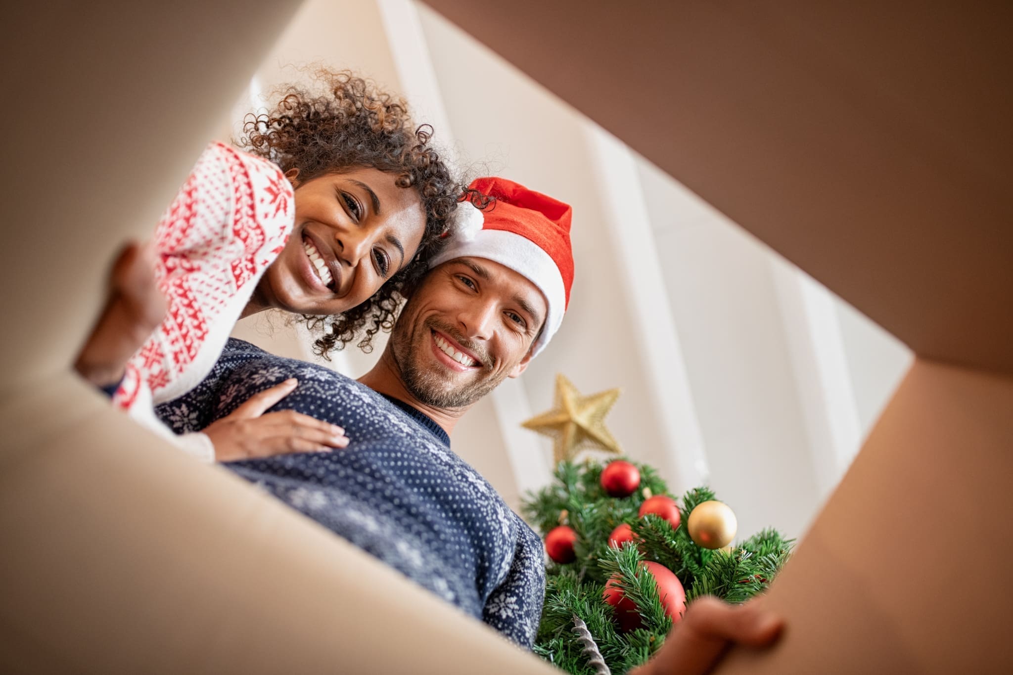 Happy multiethnic couple embracing while looking inside a gift box. Young man and beautiful african woman unpacking xmas present with christmas tree. Smiling young man wearing santa claus hat with woman looking at camera while opening present.