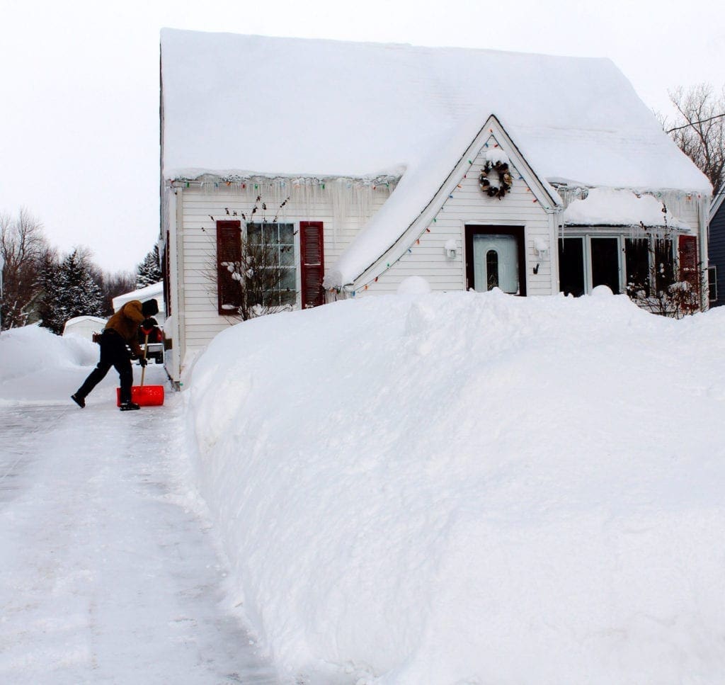 Un homme utilise une souffleuse à neige pour dégager un chemin devant une maison bleu et blanc.