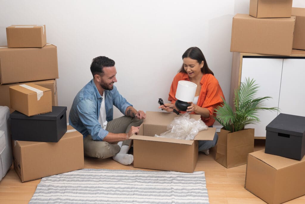Smiling couple sitting on the floor among moving boxes, unpacking home items in their new apartment.