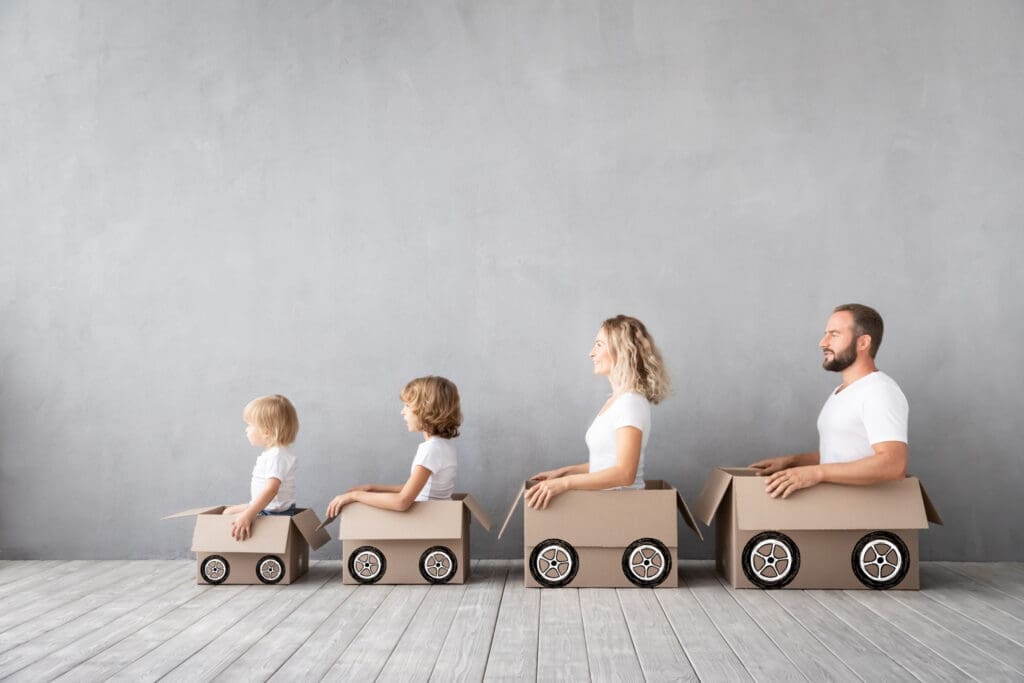 Family playfully simulates car driving with cardboard boxes as they prepare for a home move, showcasing a fun approach to transitioning kids to a new home