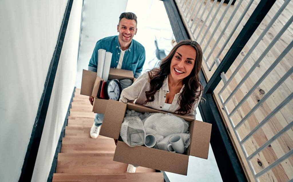 Young couple carrying a box filled with household items upstairs in their new home.