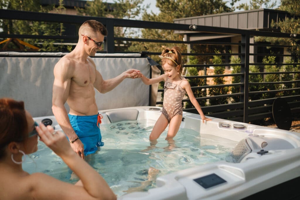 A father helps his daughter into a hot tub while a woman looks on, illustrating a family enjoying their outdoor spa.
