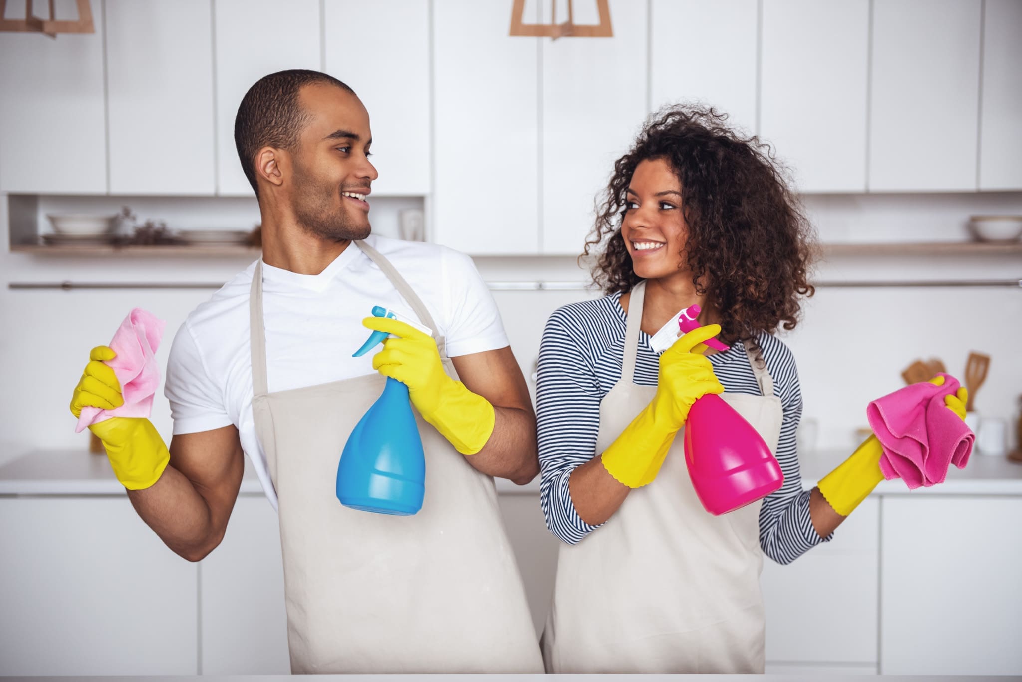 Happy couple engaging in move-out cleaning of kitchen, equipped with cleaning sprays and cloths, wearing yellow gloves.