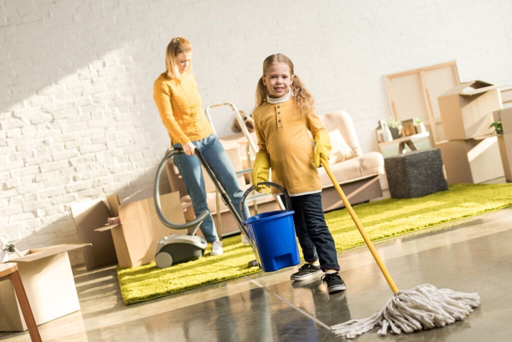 Mother vacuuming the floor while young daughter helps by mopping in a living room filled with moving boxes.