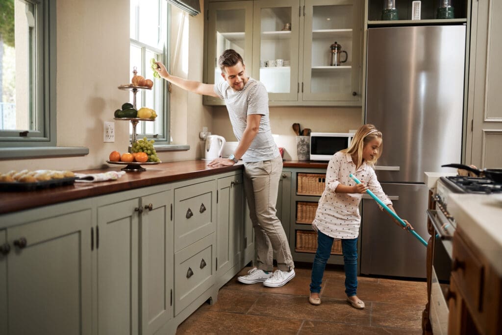 Father and daughter cleaning the kitchen together, with the daughter using a mop and the father wiping the counter.